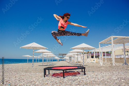 A slim athletic girl with dreadlocks jumping on a fitness trampoline outdoors on a pebble beach by the sea.