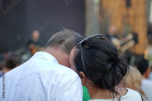 Dwoje zakochanych ludzi, mężczyzna i kobieta, w dojrzałym wieku, stoją tyłem (górne części ciała), eleganccy, pani przytula się i delikatnie całuje ukochanego, on nachyla do niej głowę, w tle ulica