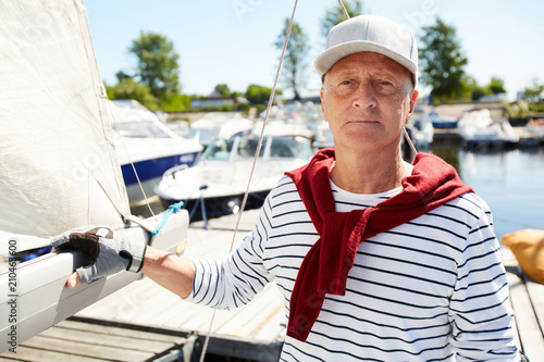 Senior active man in striped singlet and baseball cap looking at camera on hot summer day while floating on yacht