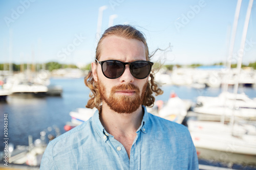 Bearded man in sunglasses and denim shirt standing in front of camera with yachts on background