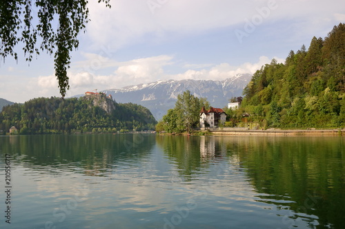 Krajobraz słoweńskiego miasteczka Bled, jezioro, promenada wzdłuż brzegu z piękną zabudową, ufortyfikowana budowla na skalistym wzniesieniu, w tle Alpy Julijskie z ośnieżonymi szczytami