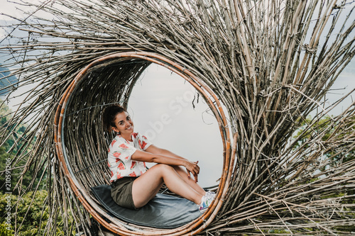 Young tourist enjoying her travel around Bali island , Indonesia. Making a stop on a beautiful hill overlooking a large lake. Photographs in a straw nest, natural environment. Lifestyle.