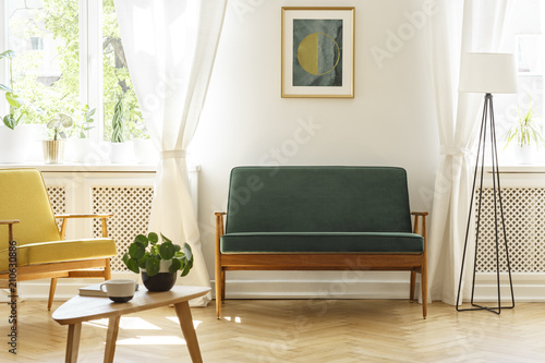 Poster above green bench between lamp and yellow armchair in vintage living room interior. Real photo