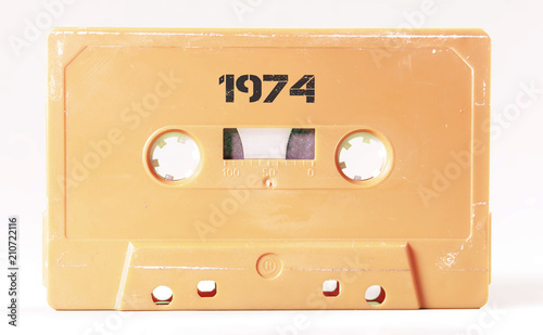 A vintage cassette tape from the 1980s era (obsolete music technology) with the text 1974 printed over it, stencil font. Color: cream, sand. White background. 