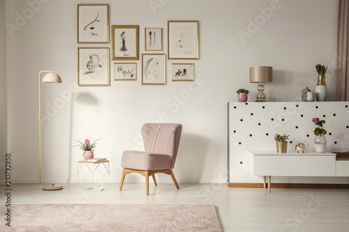 Old-fashioned armchair, pastel pink floor lamp and stylish, golden decorations in a retro living room interior with white walls