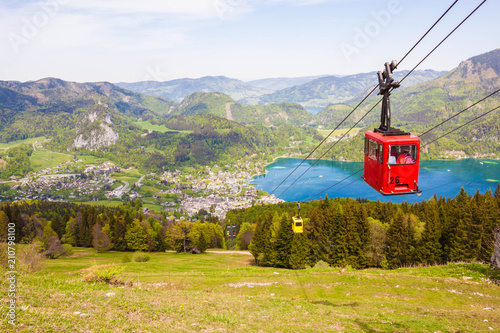 Yellow and red gondolas of Zwoelferhorn Seilbahn (cable way) travel up and down alpine peak with a view of village St.Gilgen, Wolfgangsee lake, surrounding hills and mountains. Salzburg land, Austria