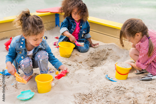 three multiethnic little children playing with plastic scoops and buckets in sandbox at playground