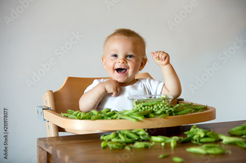 Toddler child, cute boy in white shirt, eating pea at home.