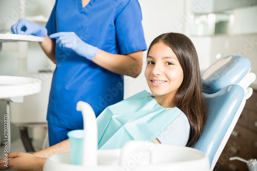 Smiling Teenager Sitting On Chair By Dentist Standing In Clinic