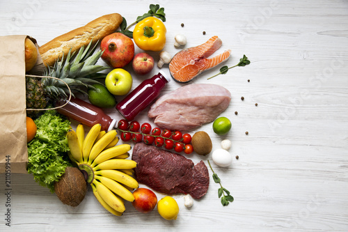 Full paper bag of healthy raw food on white wooden table. Cooking food background. Flat-lay of fresh fruits, veggies, greens, different meat, top view, copy space. Shopping concept.