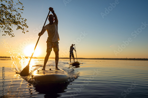 Men, friends sail on a SUP boards in a rays of rising sun