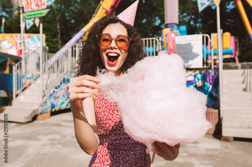 Cheerful lady with dark curly hair in sunglasses and birthday cap standing with cotton candy in hand and happily looking in camera while spending time in amusement park