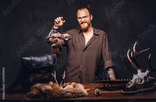 Redhead taxidermist hipster male in sunglasses dressed in a brown shirt, standing near a table with handmade trophy, owl scarecrow, and the fox skin. Isolated on the dark background.