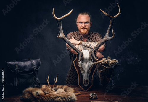 Redhead taxidermist hipster male in sunglasses dressed in a brown shirt, standing near a table with handmade trophy, owl scarecrow, and the fox skin. Isolated on the dark background.