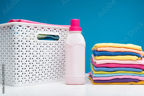 Close up of colorful garments stacked in pile. Bottle of softener and wicker clothesbasket are standing aside