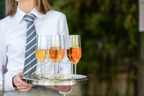 Junge Kellnerin mit weißem Hemd und Krawatte hält ein silbernes Tablett mit Champagner Gläsern in der Hand