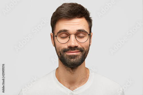 Pleased young Caucasian male closes eyes and imagines something pleasant, dreams about incredible things, wears spectacles, has dark bristle, isolated over white background. People and emotions