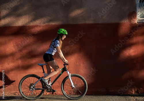 Young sporty woman in cycling clothes and helmet riding bike in front of red wall. Concept of healthy lifestyle and everyday activities. Female exercising, overcoming challanges, morning routine.