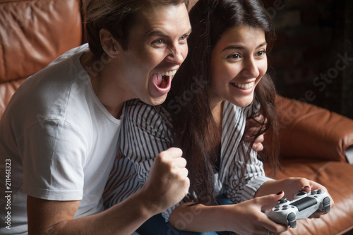 Young female gamer hold joystick, playing video game while spending weekend at home, excited boyfriend cheering her up, supporting her to win race, couple having fun together. Entertainment concept