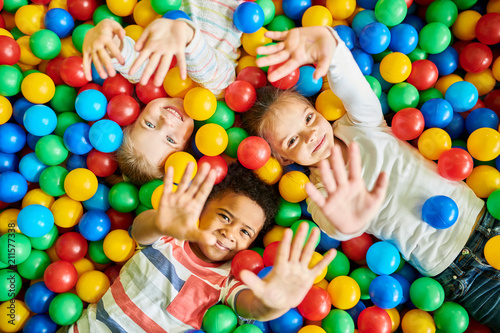 Above view portrait of three happy little kids in ball pit smiling at camera raising hands while having fun in children play center, copy space