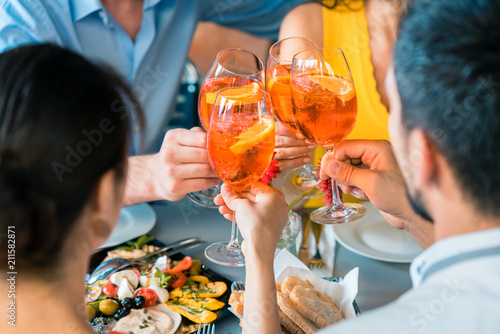 Cheerful young best friends toasting with a refreshing delicious summer drink while sitting together at table at a trendy restaurant outdoors