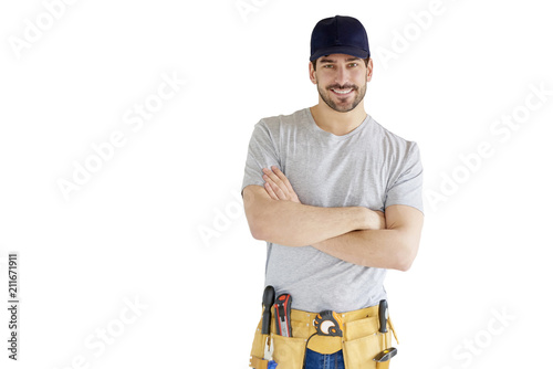 Portrait of young handyman standing at isolated white background with copy space. Successful repairman wearing baseball cap and tool belt.