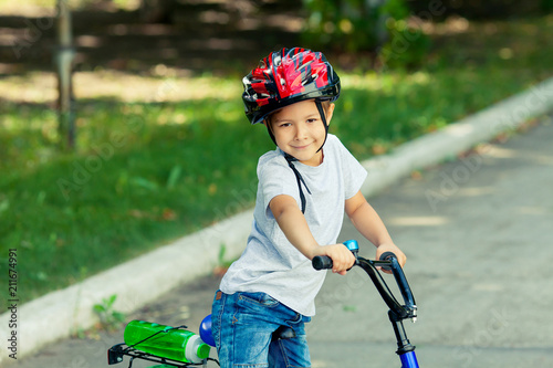 Little boy learns to ride a bike in the park near the home. Portrait of a cute kid on bicycle. Happy smiling child in helmet riding a cycling.