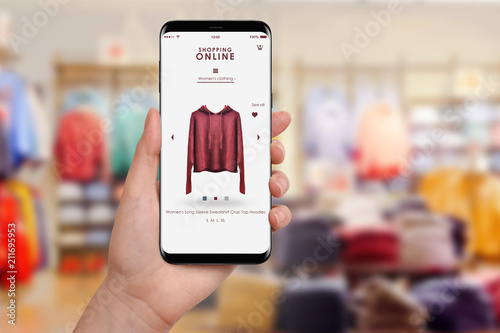 Female hand holding smartphone and shopping online, blurred clothing store in background. Online shopping concept