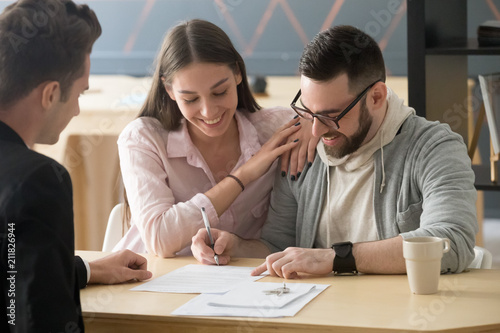 Excited millennial couple signing purchase agreement buying first home together, husband puts signature on document, becoming apartment owner, spouses legalize property ownership in realtor office