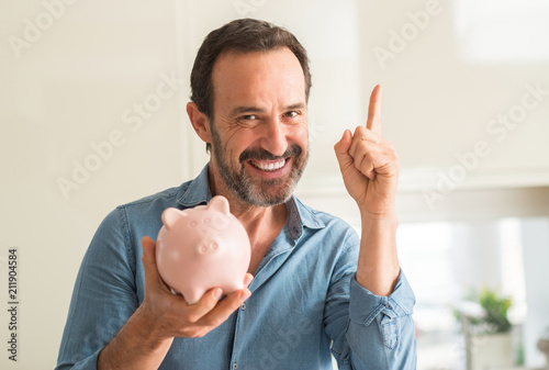 Middle age man save money on piggy bank surprised with an idea or question pointing finger with happy face, number one