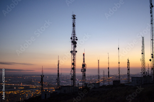 communication tower. cell, radio and television antennas on top of a mountain and below a lit coastal village