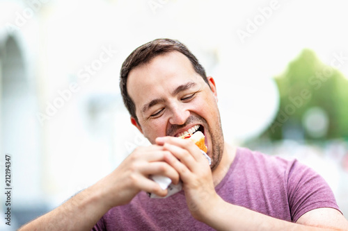 Closeup portrait of hungry man biting hot dog at city street background.