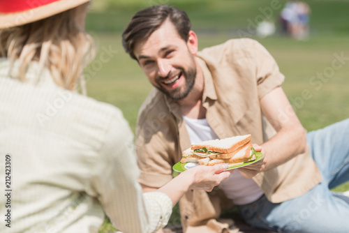 young couple holding sandwiches while spending time together at picnic in park