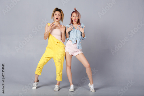 Full length of two cheerful young female in color casual demin clothes having shocked and scared expressions, mouths wide open, feeling nervous and frightened. People emotions
