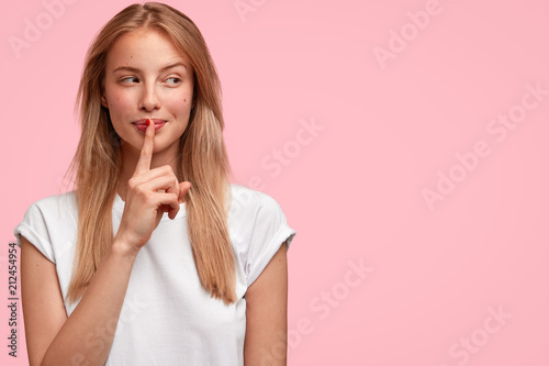 Indoor shot of beautiful female looks mysteriously aside, has intriguing look, asks to be quiet, dressed in casual clothes, stands against pink background with blank copy space for your advertisement