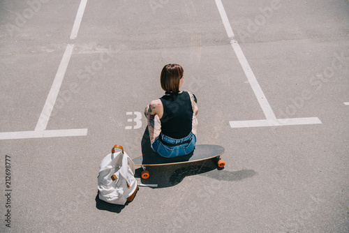 rear view of stylish tattooed girl sitting on skateboard near backpack at parking lot