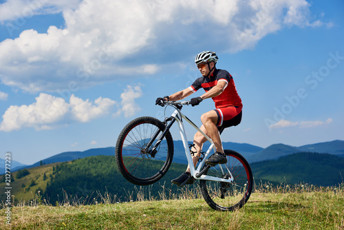 Athletic sportsman cyclist in professional sportswear and helmet riding cross country bike, turning on one wheel of bicycle on mountain hill. Blue summer sky on background. Extreme sport concept