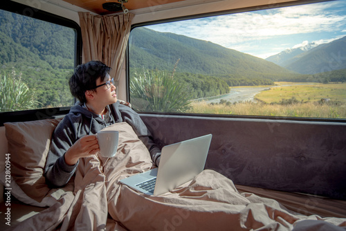 Young Asian man drinking hot coffee working with laptop computer on the bed in camper van with mountain scenic view through the window, digital nomad on road trip concept