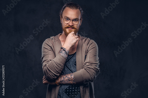 Smart pensive redhead hipster with full beard and glasses dressed in casual clothes, poses with hand on chin in a studio. Isolated on a dark background.