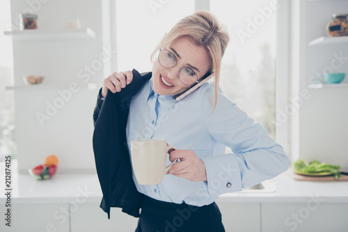 Young attractive adorable smiling blonde woman office executive worker wearing spectacles in hurry early in the morning talking on the phone putting dark blue jacket on having a drink in kitchen