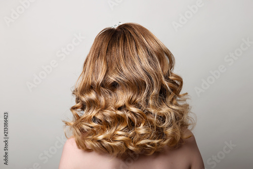 Female hairstyle with long curls on the head of a blonde with a back view on a gray background.
