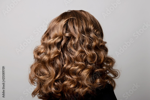 Female hairstyle long curls on the head of the brown-haired woman back view at the gray background turning the head to the right.
