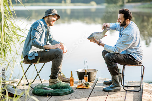 Two friends sitting together with beer and fish on the picnic while fishing near the lake