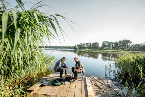 Two fishermen relaxing during the picnic on the beautiful wooden pier with green reed on the lake in the morning