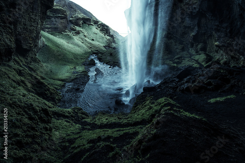 A wild natural fresh sparkling waterfall in a lonely green valley with moos covered rocks and ice cold mountain water flowing down black rocky walls on a rainy moody day in Iceland without tourists