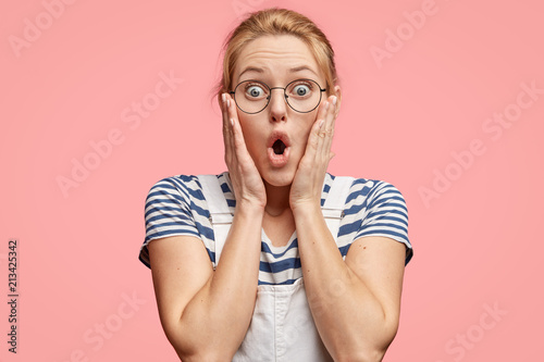 Indoor shot of pretty female with astonished expression, keeps hands on cheeks, stares at camera, being frightened of recieving bills with high sums, feels embarrassed, stands over pink background