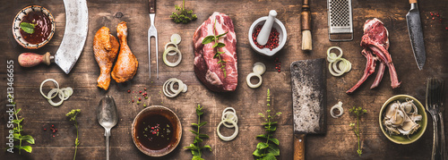 Flat lay of various grill and bbq meat : chicken legs, steaks, lamb ribs with vintage kitchenware kitchen utensils: Meat Fork and Butcher Cleaver and herbs knife. Sauces and ingredients for grilling,