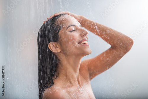 Side view beaming lady locating under stream of water. She holding hair with arms