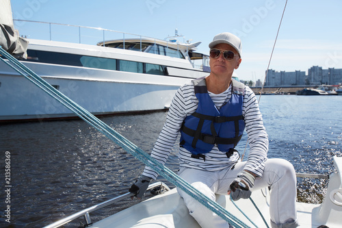 Serious pensive handsome mature male yacht captain in sunglasses and white cap wearing life jacket sitting on boat deck and looking into distance