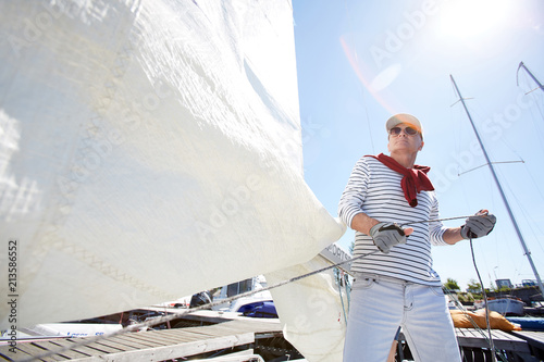 Serious mature sailor in sunglasses and cap wearing sweater wrapped around neck pulling rope while releasing sail in sunlight outdoors